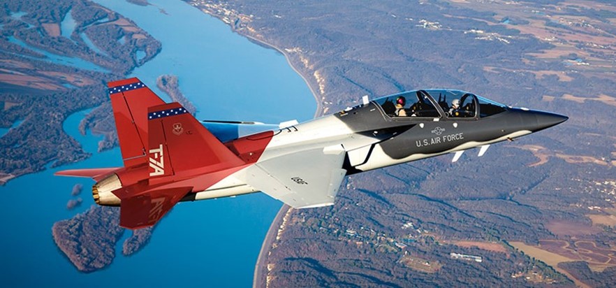 A red, white and blue T-7A Red Hawk trainer jet with two pilots in the cockpit, flying over a river and farmland